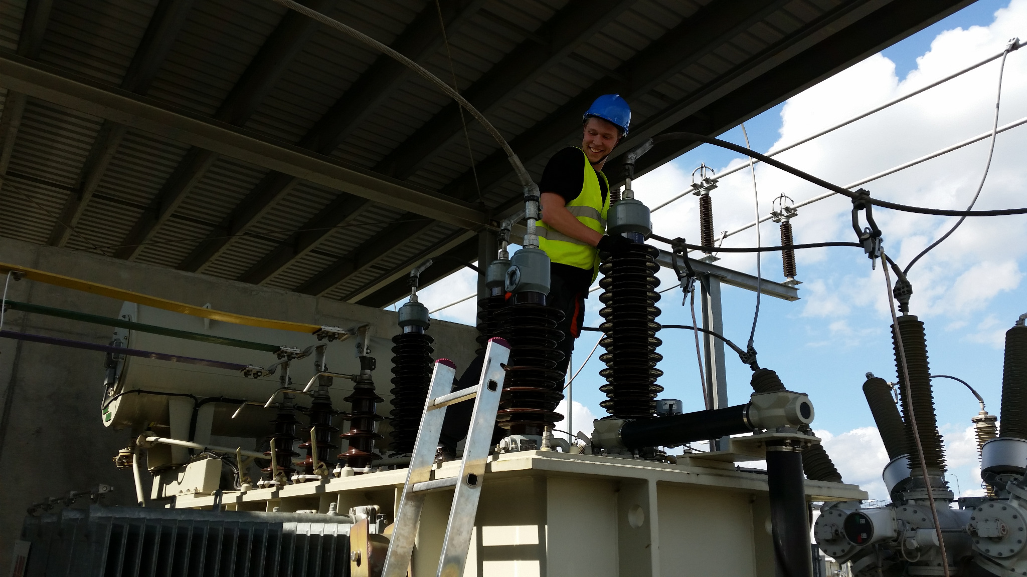 Uśmiechnięty elektryk w kasku i kamizelce na drabinie, pracujący przy transformatorze wysokiego napięcia z ceramicznymi izolatorami pod zadaszeniem, widoczne przewody i słupy energetyczne w tle.