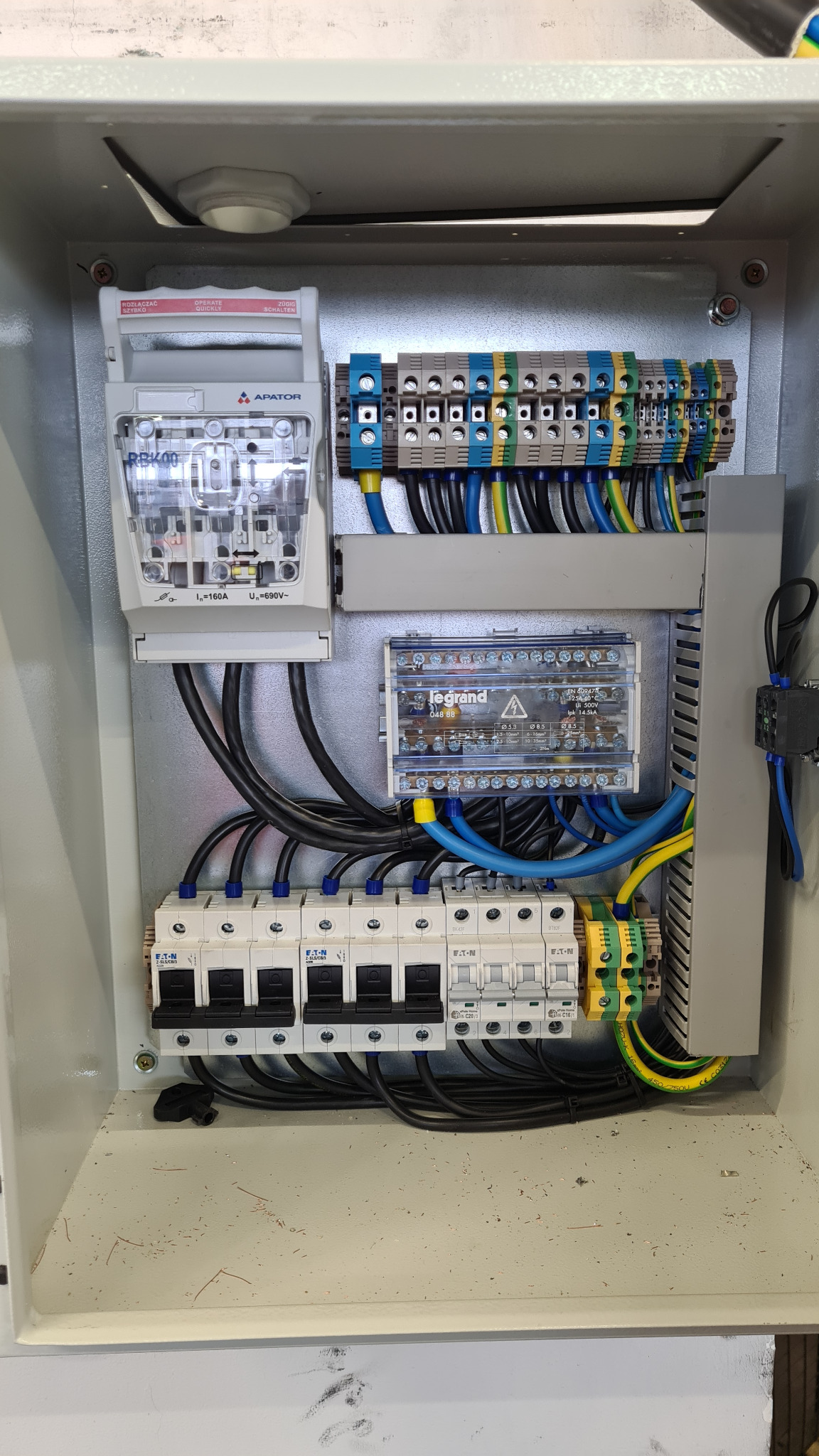 Wnętrze skrzynki elektrycznej z zamontowanym licznikiem Apator, zabezpieczeniami Eaton, modułem Legrand i listwami zaciskowymi z przewodami w kolorach niebieskim, czarnym, żółto-zielonym.