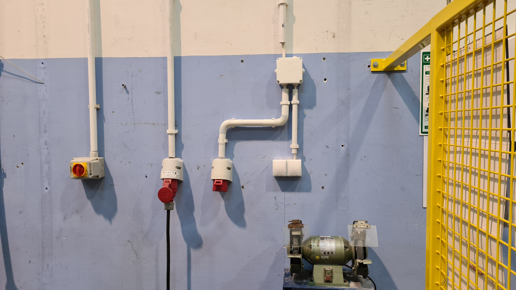 Ściana warsztatu z widocznymi elementami instalacji elektrycznej: gniazda przemysłowe z czerwonymi wtykami, wyłącznik obrotowy, puszka instalacyjna z peszlem, podwójny włącznik; na dole szlifierka...
