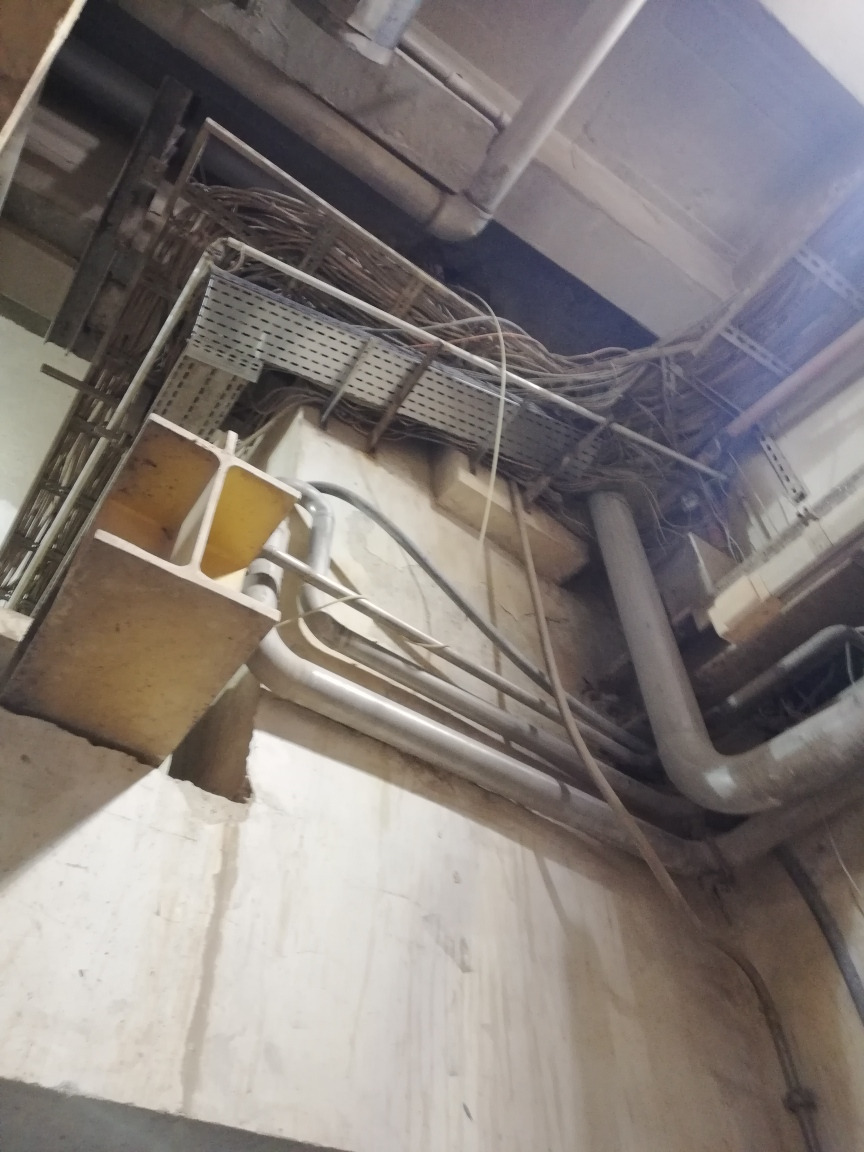 Widok z dołu na koryta kablowe, rury i przewody elektryczne zamontowane pod sufitem w pomieszczeniu technicznym. Widoczne mocowania do ściany i sufitu, oraz liczne okablowanie.