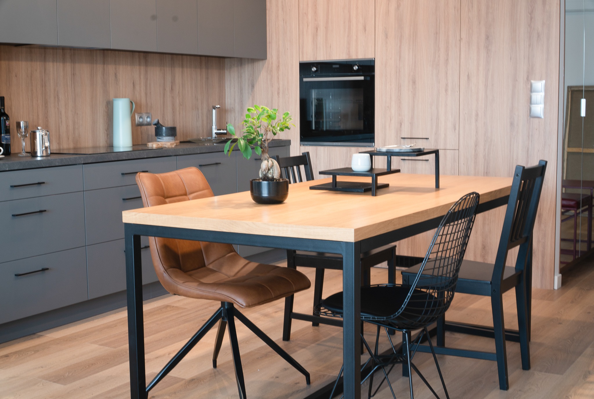 Jasny drewniany stół z czarną metalową ramą w nowoczesnej kuchni, otoczony brązowym skórzanym krzesłem obrotowym i czarnymi krzesłami, z szafkami w odcieniach szarości i drewna w tle.