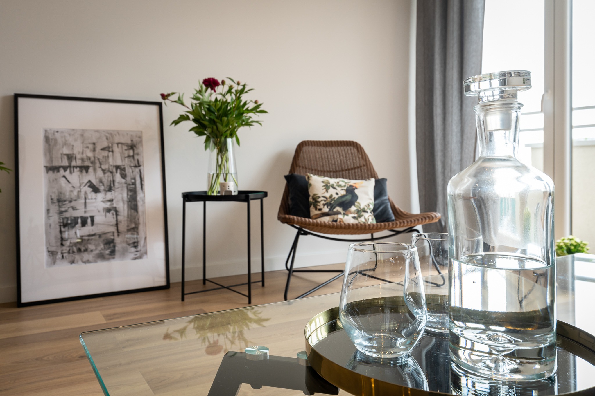 Jasne wnętrze z drewnianą podłogą, szklanym stolikiem kawowym z złotą tacą, karafką i dwoma szklankami, rattanowym krzesłem z poduszkami, stolikiem z kwiatami i abstrakcyjnym obrazem w czarnej ramie.