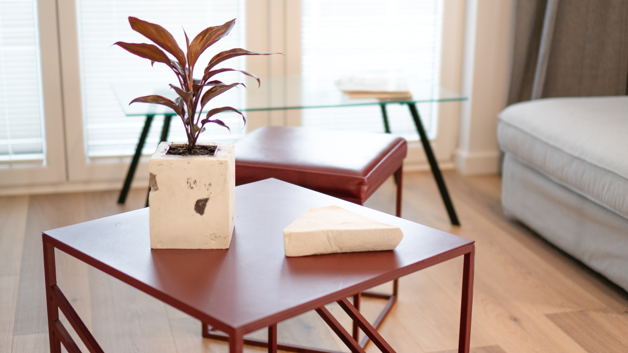 Nowoczesny salon z niskim stolikiem kawowym w kolorze burgundu, na którym stoi betonowa doniczka z rośliną i dekoracyjny element w kształcie trójkąta, w tle szklany stolik i fragment sofy.