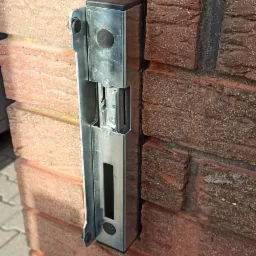 Elektrozaczep zamontowany na ceglanym słupku ogrodzenia, widoczne ślady po wierceniu i montażu.