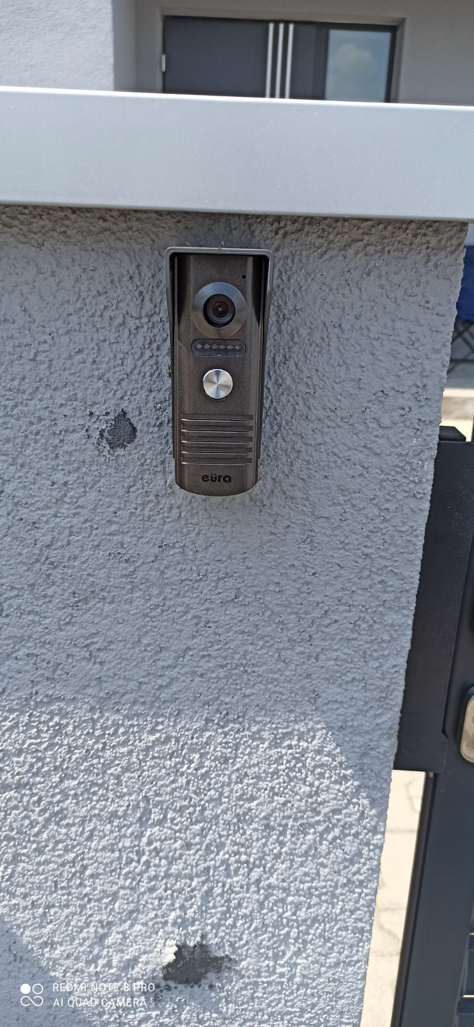 Szary wideodomofon Eura zamontowany na teksturowanej, jasnoszarej ścianie przy wejściu, widoczne elementy bramy po prawej stronie.