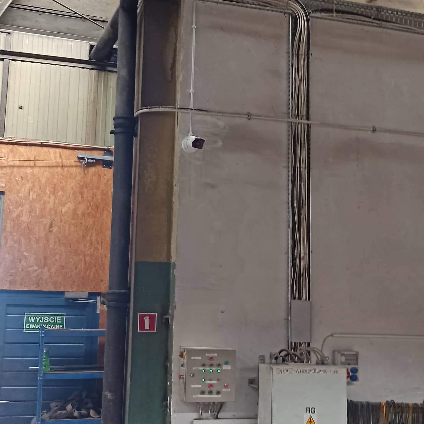 Wnętrze hali produkcyjnej z widoczną kamerą przemysłową na ścianie, okablowaniem w korytkach, tablicą sterowniczą i znakiem ewakuacyjnym.