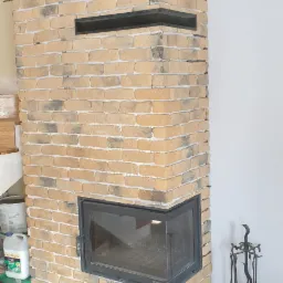 Obudowa kominka z cegły klinkierowej w odcieniach beżu i szarości, z czarnymi wnękami i metalowym wkładem kominkowym, na podłodze wyłożonej płytkami imitującymi drewno; widoczne akcesoria kominkowe.