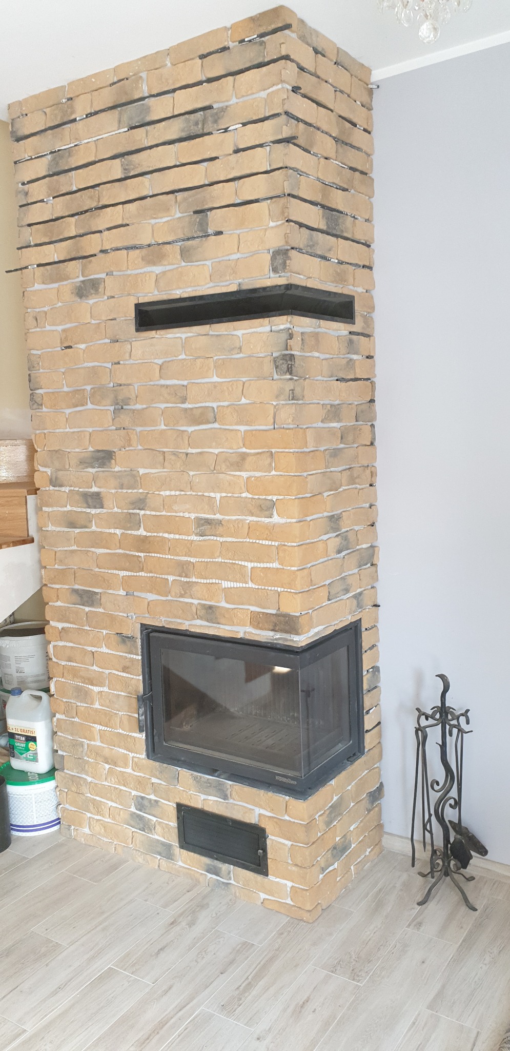 Obudowa kominka z cegły klinkierowej w odcieniach beżu i szarości, z czarnymi wnękami i metalowym wkładem kominkowym, na podłodze wyłożonej płytkami imitującymi drewno; widoczne akcesoria kominkowe.