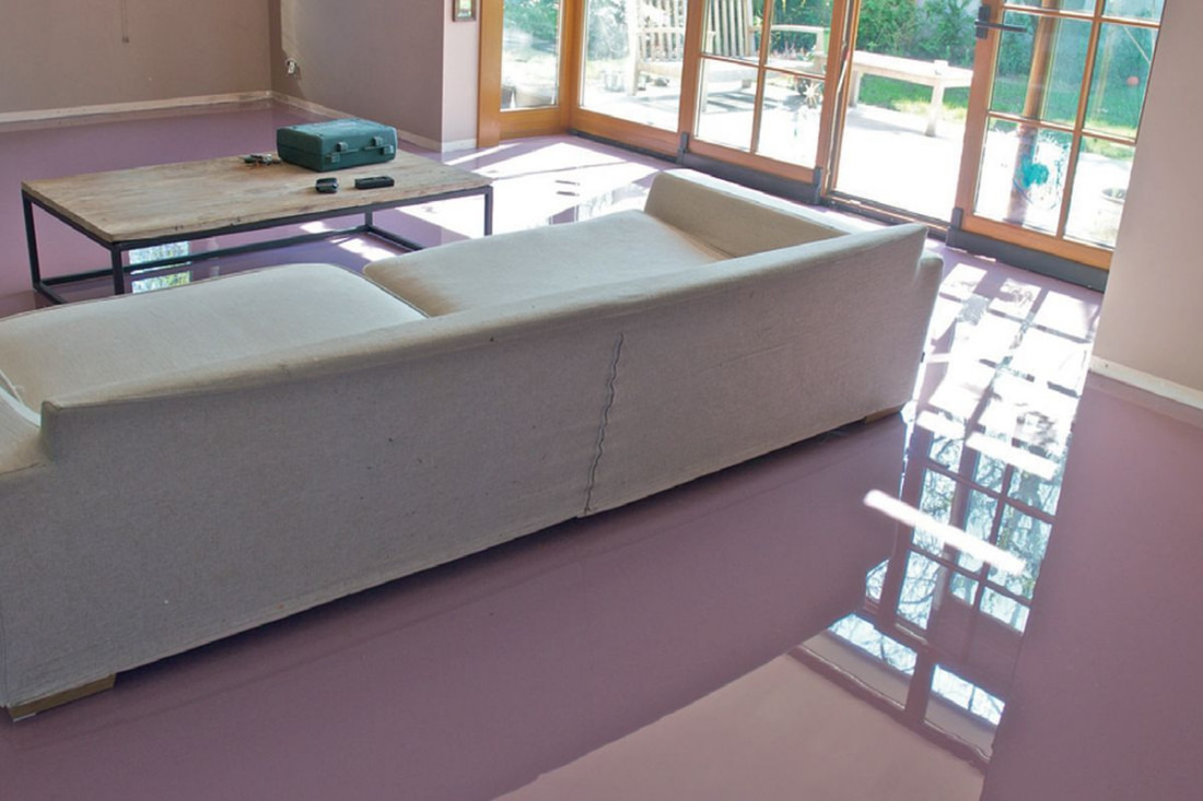 Salon z błyszczącą, jednolitą podłogą w odcieniach różu, odbijającą światło z okien balkonowych, z widoczną sofą i stolikiem kawowym z narzędziami w tle.