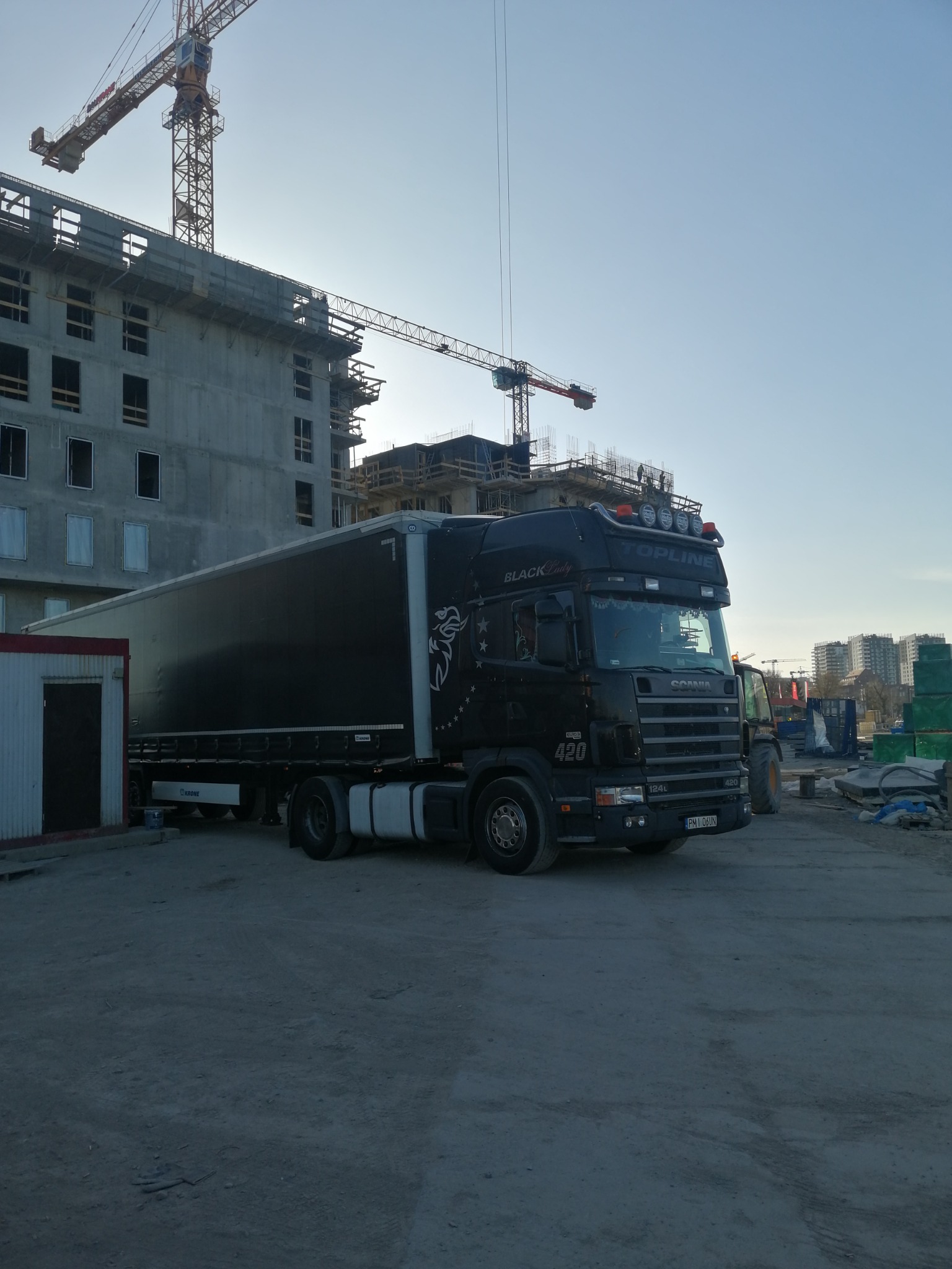 Czarna ciężarówka Scania Topline z naczepą Krone, z grafiką pegaza i napisem Black Lady, zaparkowana na placu budowy z dźwigami w tle.