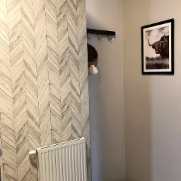 Przedpokój z tapetą w jodełkę imitującą drewno, białym grzejnikiem, czarnym wieszakiem z koszykiem i obrazem szkockiej krowy w czarnej ramie.