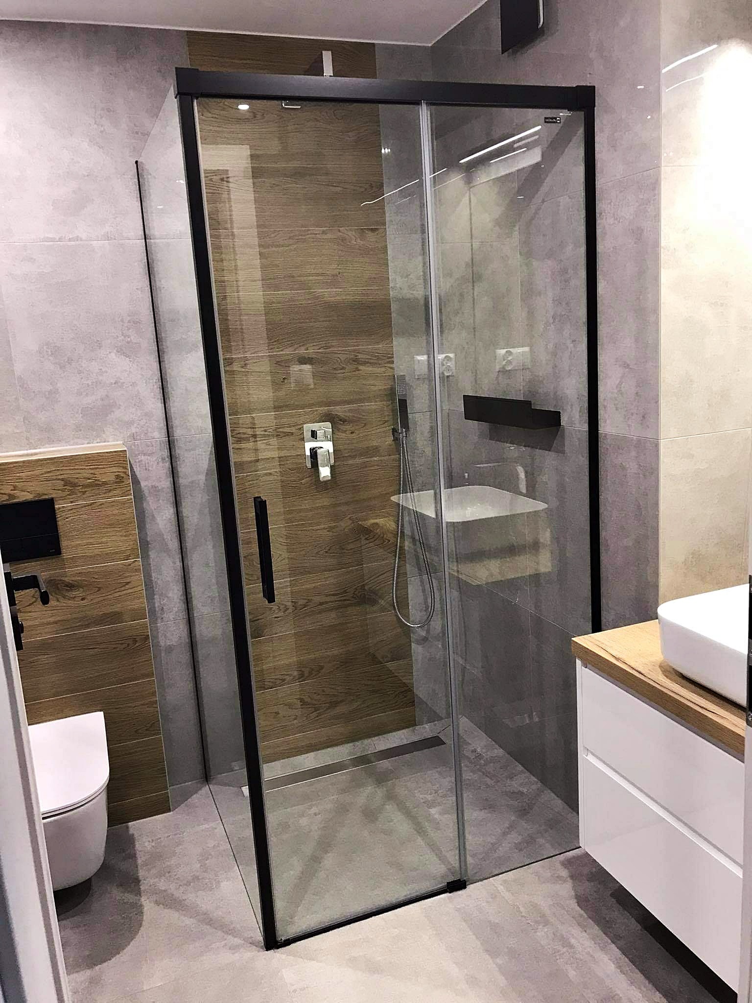 Nowoczesna łazienka z kabiną prysznicową typu walk-in z czarnymi ramami, drewnopodobnymi płytkami na ścianie wewnątrz kabiny i szarymi płytkami na pozostałych ścianach, biała umywalka nablatowa...
