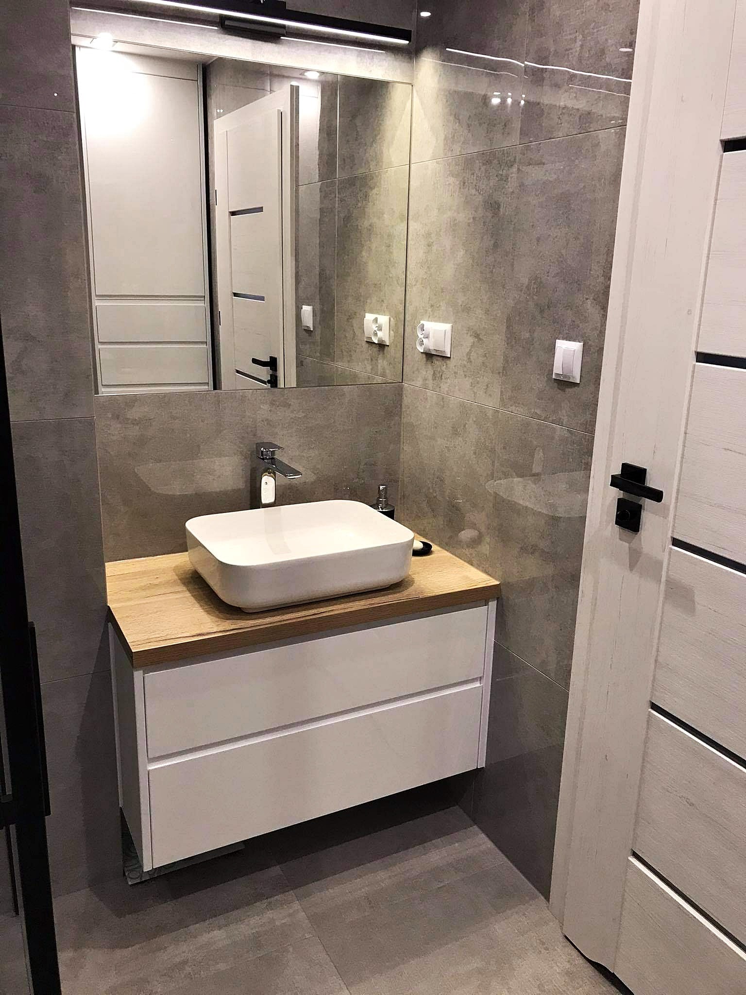 Nowoczesna łazienka z szarymi płytkami imitującymi beton, białą szafką pod umywalką z drewnianym blatem, prostokątną umywalką nablatową i dużym lustrem.