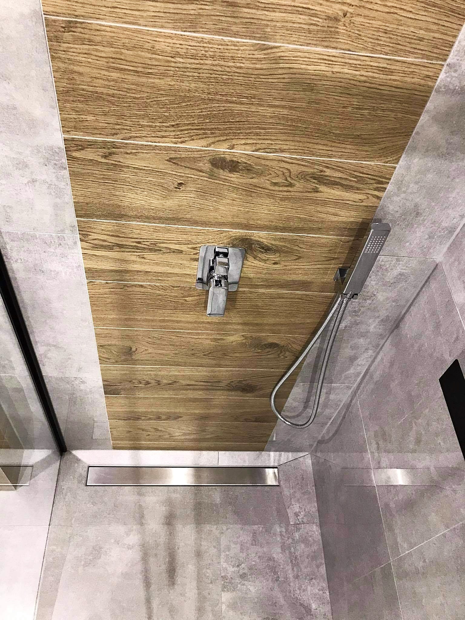 Nowoczesna kabina prysznicowa z drewnopodobnymi płytkami na suficie, szare płytki na ścianach, bateria podtynkowa i odpływ liniowy w podłodze.