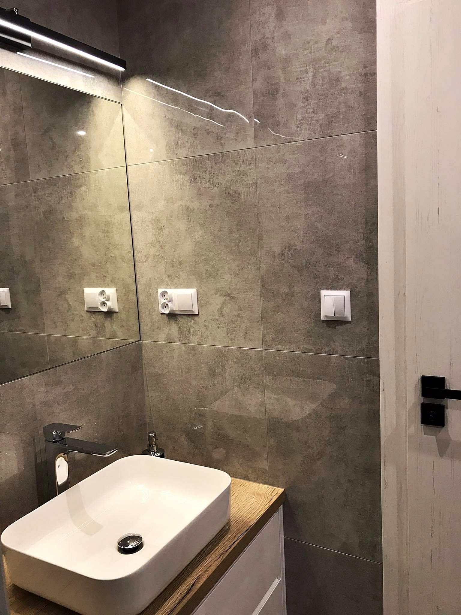 Nowoczesna łazienka z umywalką nablatową, chromowaną baterią i szarymi płytkami imitującymi beton, oświetlenie LED nad lustrem.