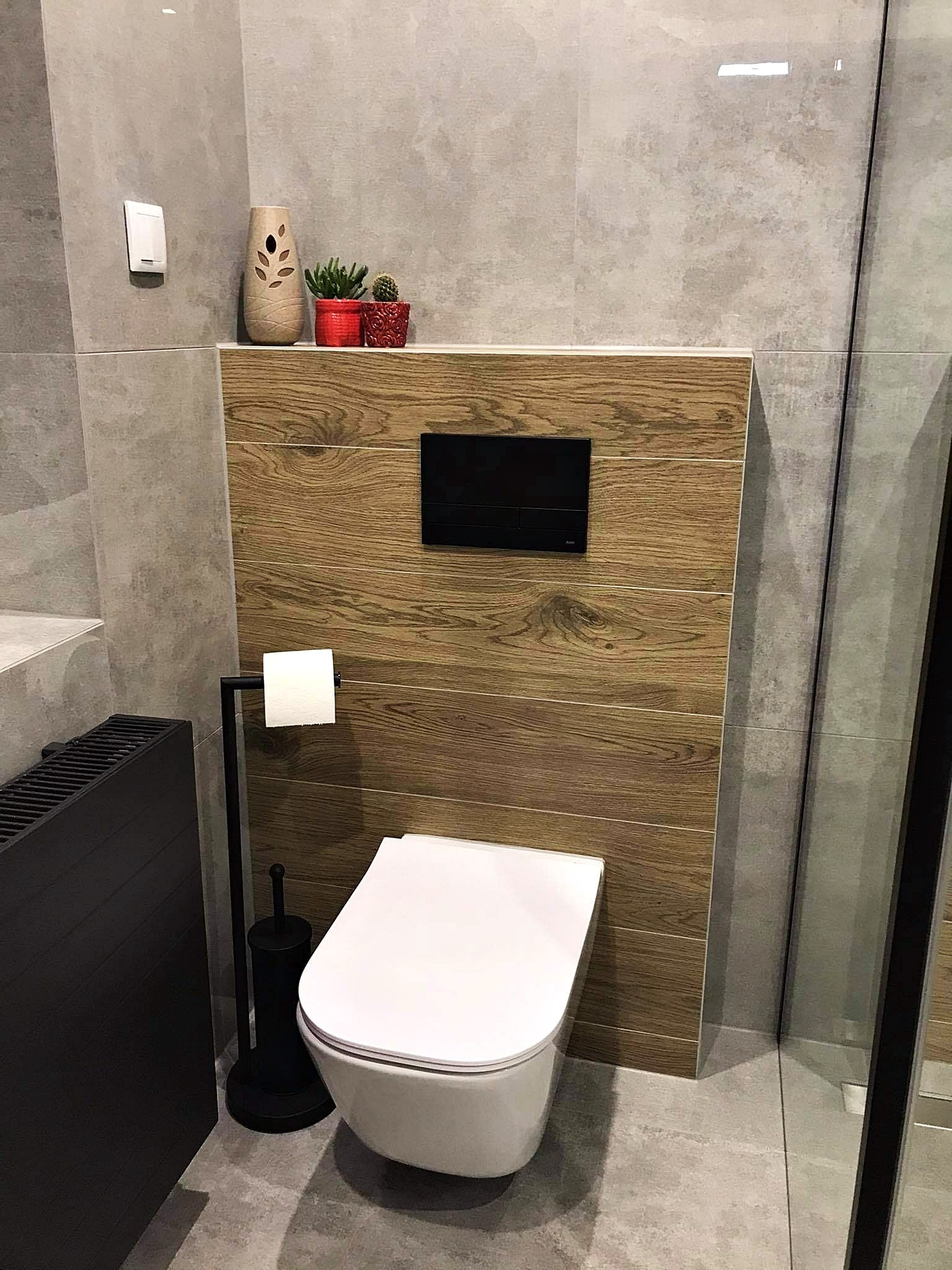 Nowoczesna toaleta podwieszana z drewnianym panelem, czarnym przyciskiem spłukującym, grzejnikiem i akcesoriami w szaro-beżowej łazience.