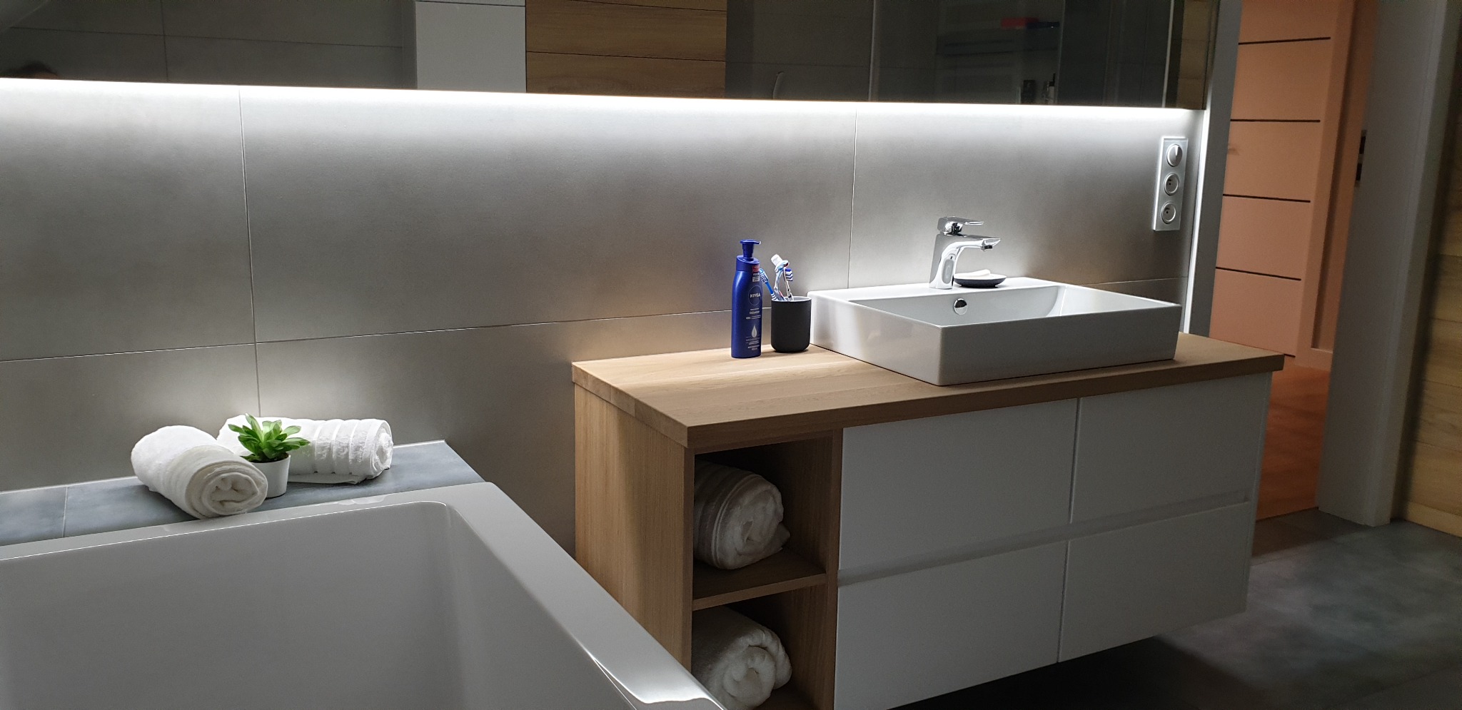 Nowoczesna łazienka z białą umywalką nablatową, drewnianym blatem i szafką z szufladami, podświetlane lustro, szare płytki i dekoracyjne ręczniki.