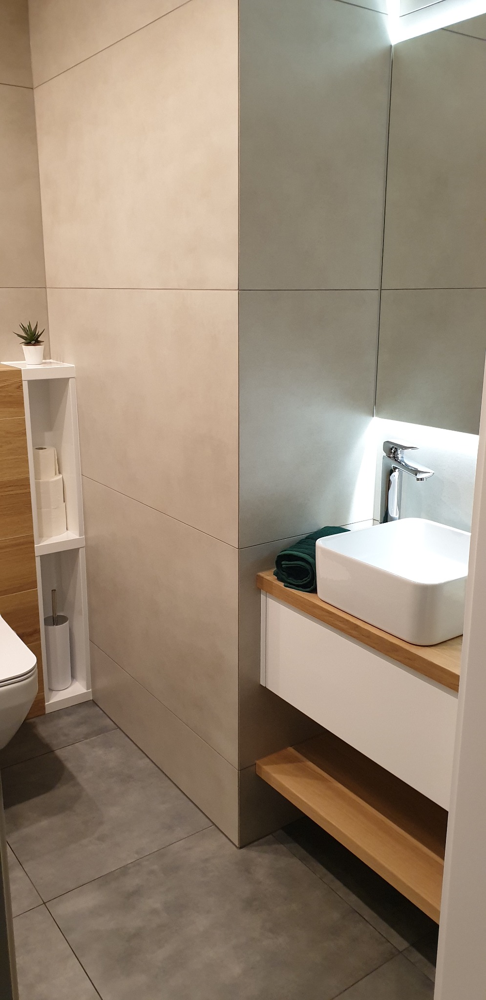 Nowoczesna łazienka z kwadratową umywalką nablatową, drewnianym blatem i półką, szarą podłogą i ścianami z płytek w odcieniach beżu i szarości, z akcentami drewna i rośliną doniczkową.