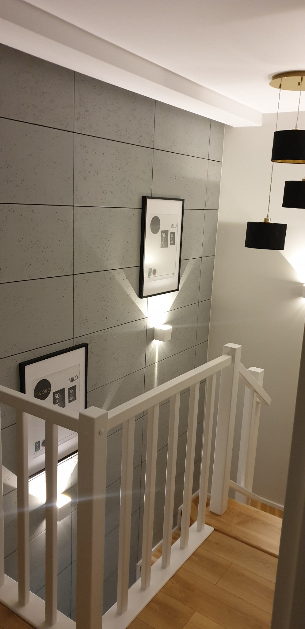 Nowoczesne wnętrze z szarą ścianą wyłożoną kwadratowymi płytami, białymi schodami z drewnianymi stopniami i czarnymi lampami wiszącymi