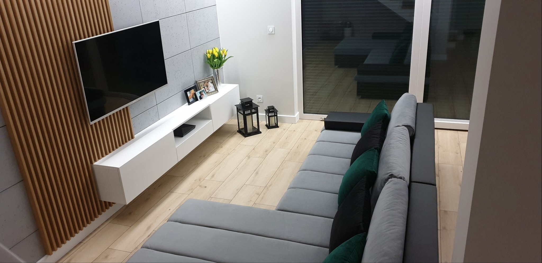 Nowoczesny salon z szarym narożnikiem, drewnianymi lamelami na ścianie za telewizorem, białymi meblami i drewnianą podłogą, doświetlony przez duże okno balkonowe.