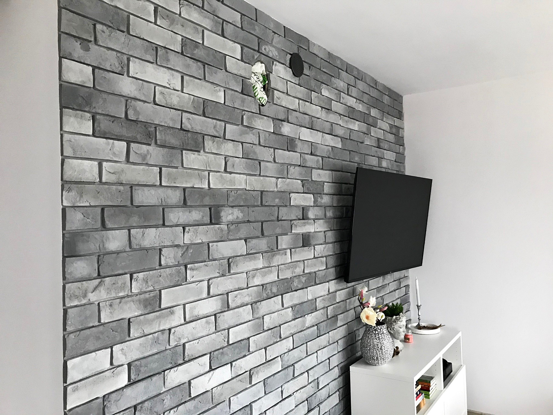 Ściana wykończona szarą cegłą dekoracyjną, z ukrytym kablem i zamontowanym telewizorem, uzupełniona białym regałem z dekoracjami.