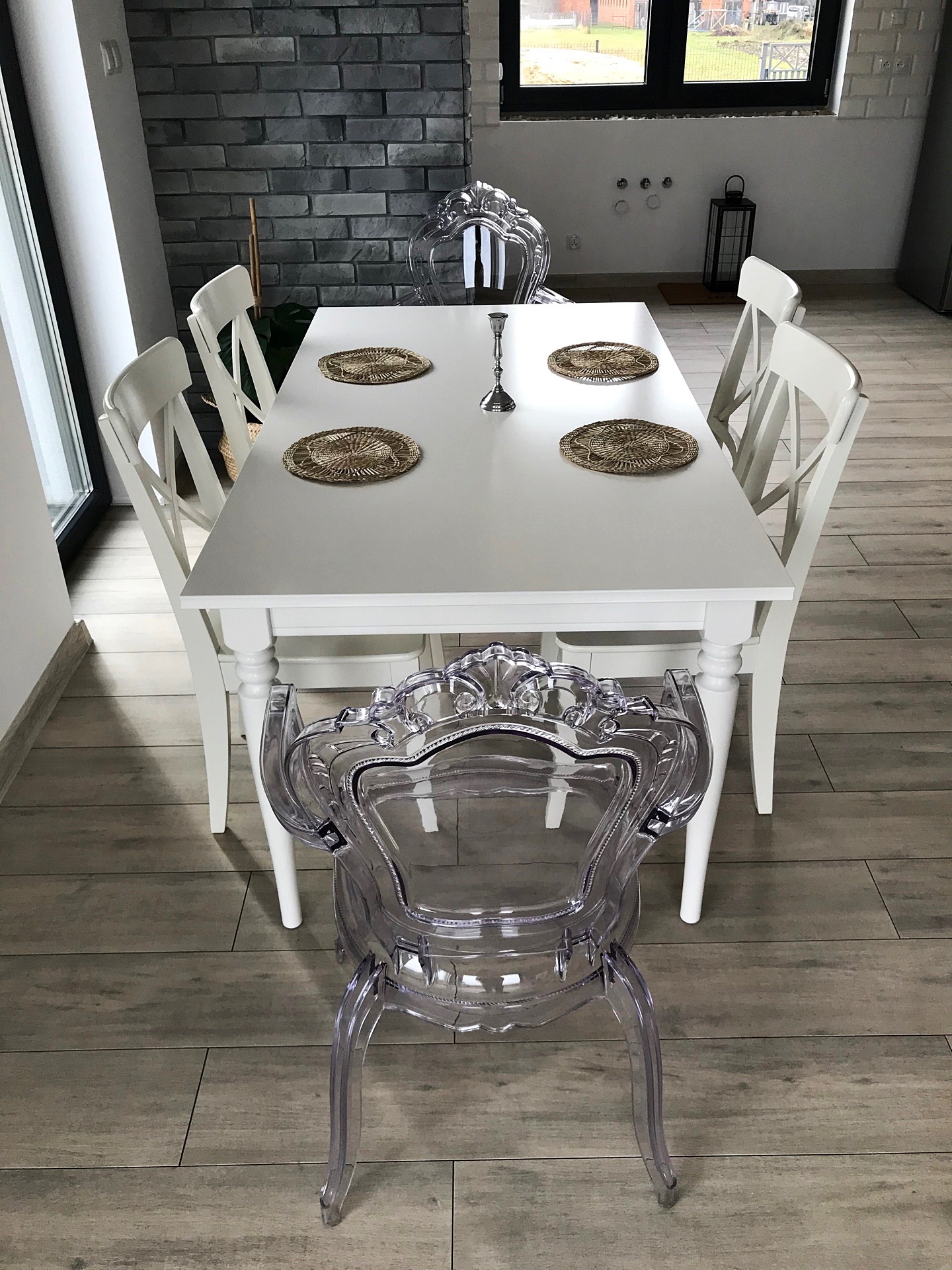 Jadalnia z białym stołem otoczonym białymi krzesłami i krzesłami z przezroczystego tworzywa, dekoracyjne podkładki na stole, szara ściana z cegły w tle.
