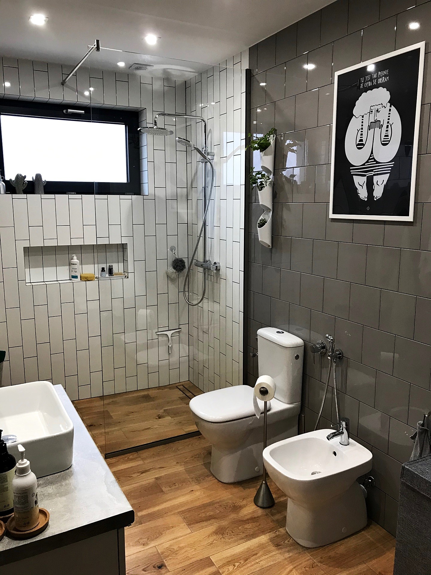 Nowoczesna łazienka z drewnianą podłogą, białą armaturą i szarymi płytkami, ozdobiona minimalistycznym plakatem i roślinami w wiszących doniczkach.