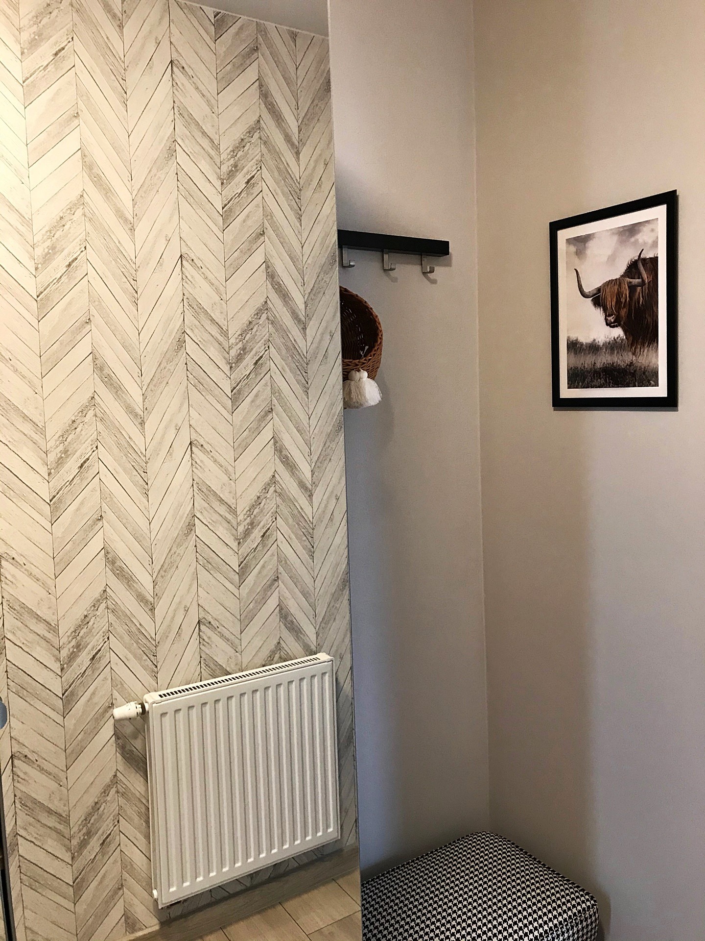 Przedpokój z tapetą w jodełkę imitującą drewno, białym grzejnikiem, czarnym wieszakiem z koszykiem i obrazem szkockiej krowy w czarnej ramie.