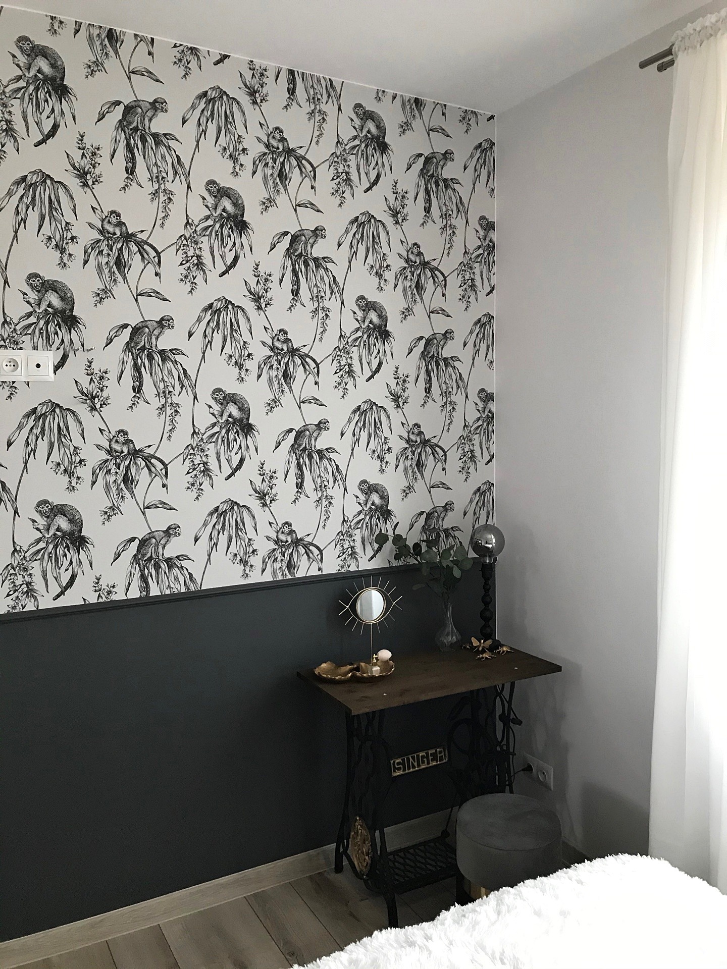 Ściana w pokoju podzielona na dwie części: górna pokryta tapetą w czarno-białe małpy i rośliny, dolna pomalowana na ciemny szary kolor. Obok ściany stoi stolik ze starą maszyną do szycia Singer...