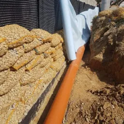 Fundament izolowany czarną folią kubełkową, z drenażem z rury pomarańczowej i otuliną z białej geowłókniny, zabezpieczony workami z kamieniami.