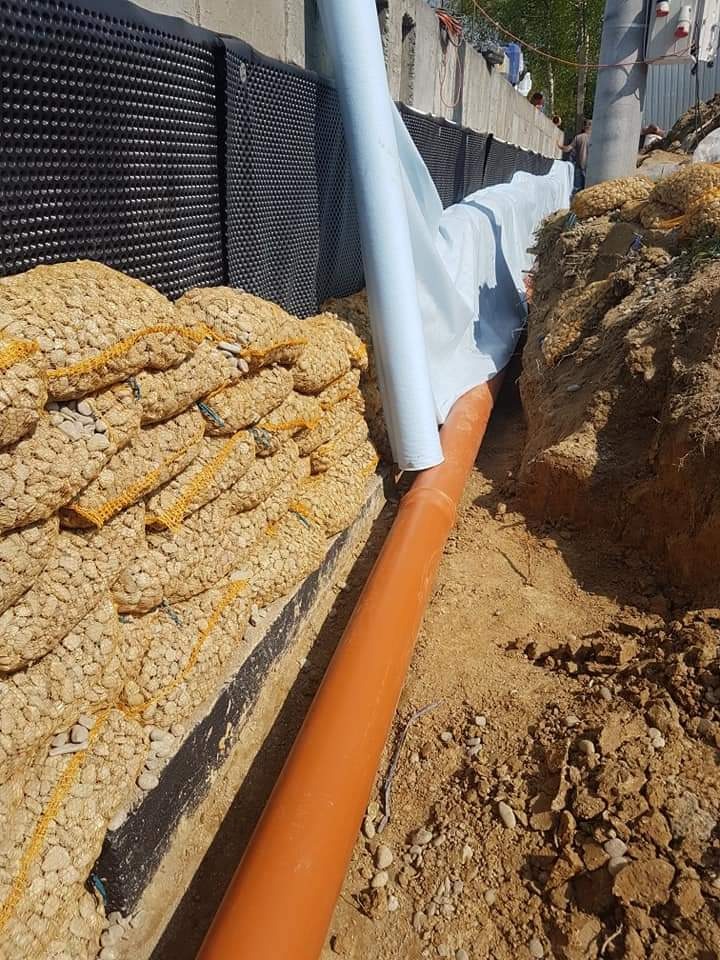 Fundament izolowany czarną folią kubełkową, z drenażem z rury pomarańczowej i otuliną z białej geowłókniny, zabezpieczony workami z kamieniami.