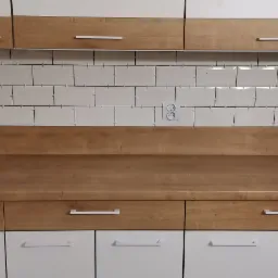 Kuchnia po remoncie: Białe, prostokątne płytki na ścianie z widocznymi krzyżykami dystansowymi, drewniany blat i szafki z białymi frontami i podłużnymi uchwytami, ż&oacute;łta miarka na blacie.