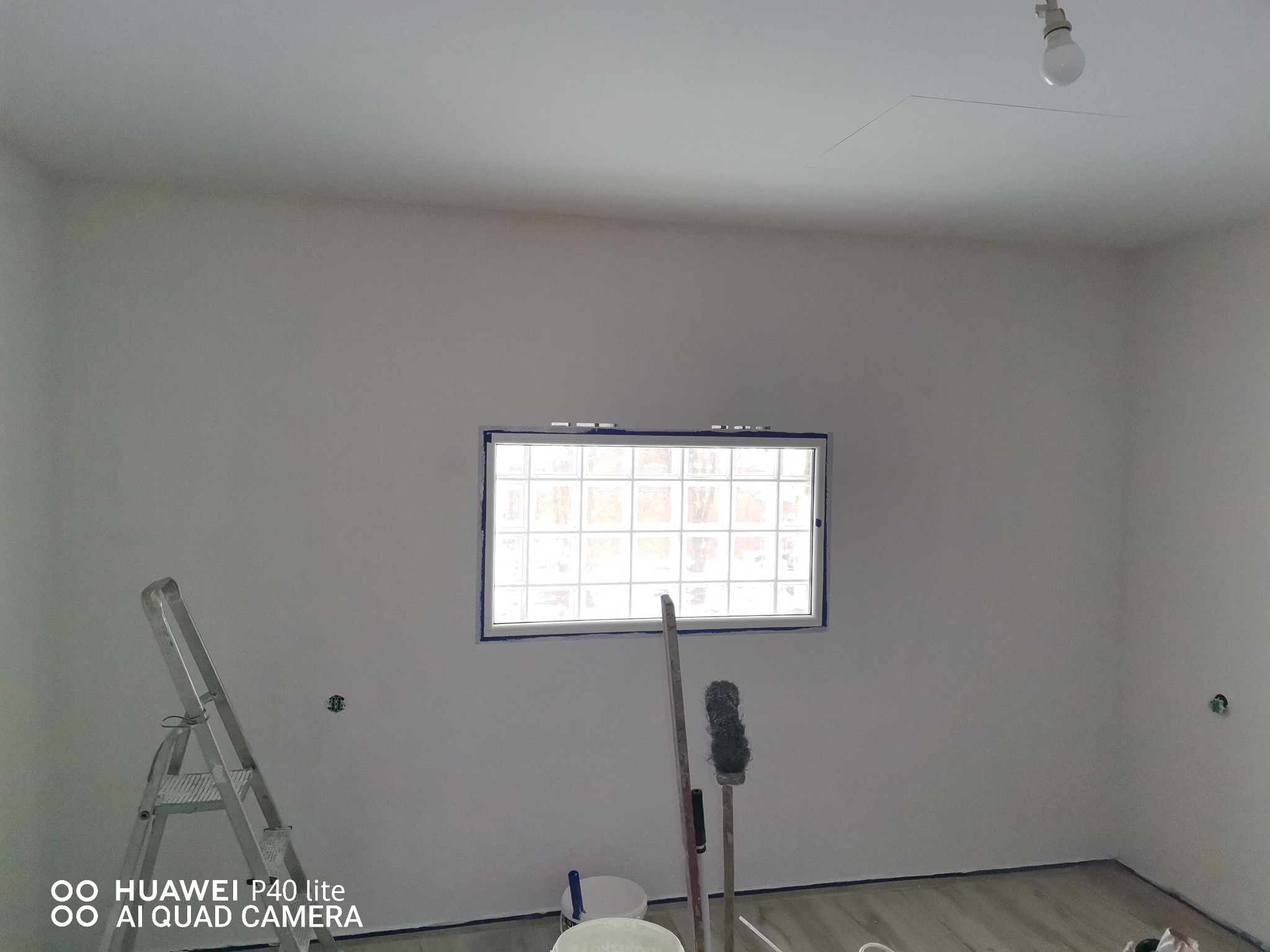 Pokój w trakcie odświeżania: ściany po nałożeniu pierwszej warstwy farby, zabezpieczone taśmą okno z luksferami, drabina malarska, wałki i pędzle w wiadrach, widoczny fragment podłogi.