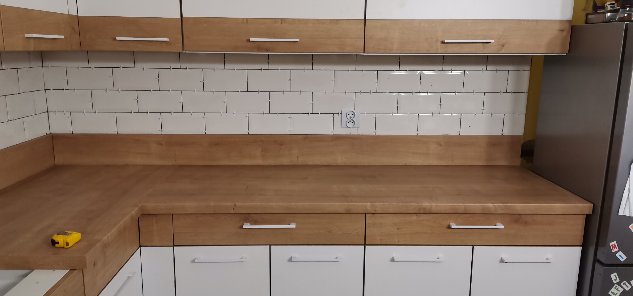Kuchnia po remoncie: Białe, prostokątne płytki na ścianie z widocznymi krzyżykami dystansowymi, drewniany blat i szafki z białymi frontami i podłużnymi uchwytami, żółta miarka na blacie.
