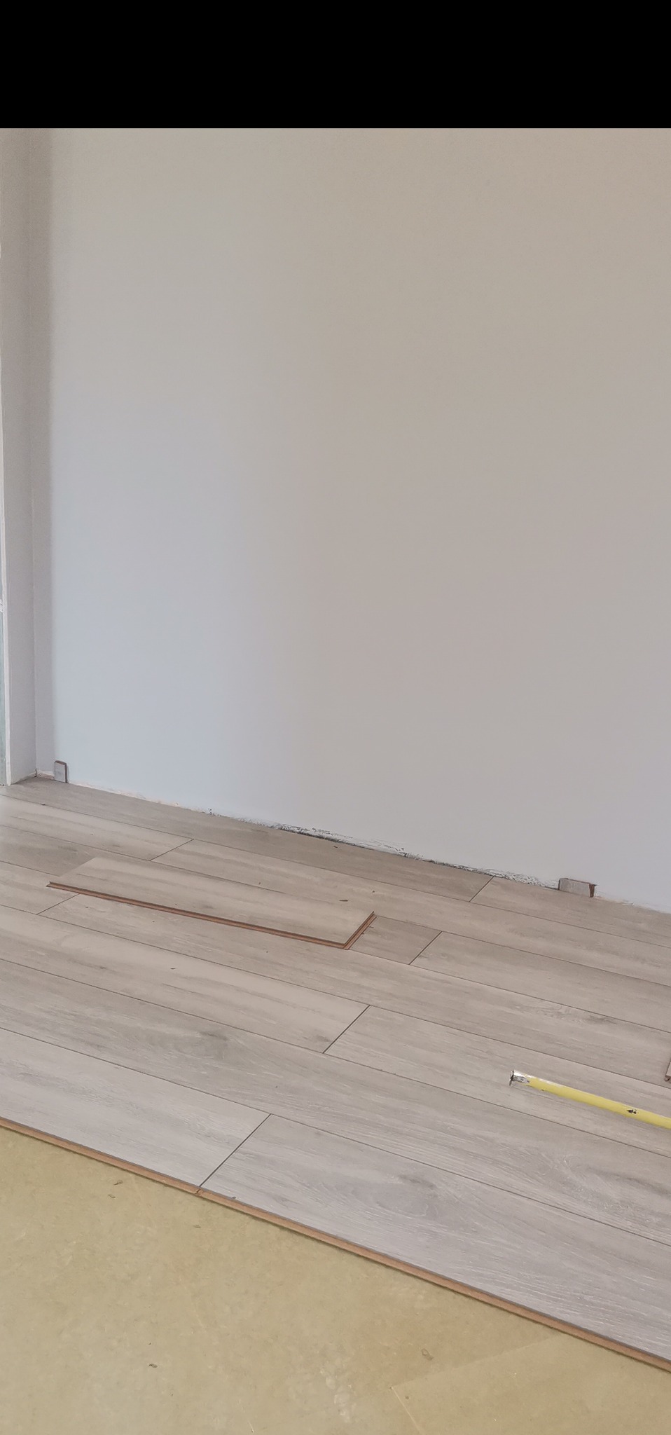 Fragment podłogi w trakcie układania paneli laminowanych o jasnym, drewnopodobnym wzorze; widoczna żółta miara zwinięta na podłodze i niedokończony rząd przy ścianie.