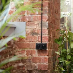 Czarna podwójna puszka elektryczna zamontowana na ceglanej ścianie, z kablem biegnącym wzdłuż ściany, widoczne rośliny doniczkowe i fragment niebieskiego pufa.