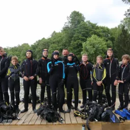 Grupa dzieci i dorosłych w czarnych piankach do nurkowania, stojąca na drewnianym pomoście nad jeziorem, z zielonym lasem w tle i sprzętem do nurkowania u stóp, w tym płetwy i butle.