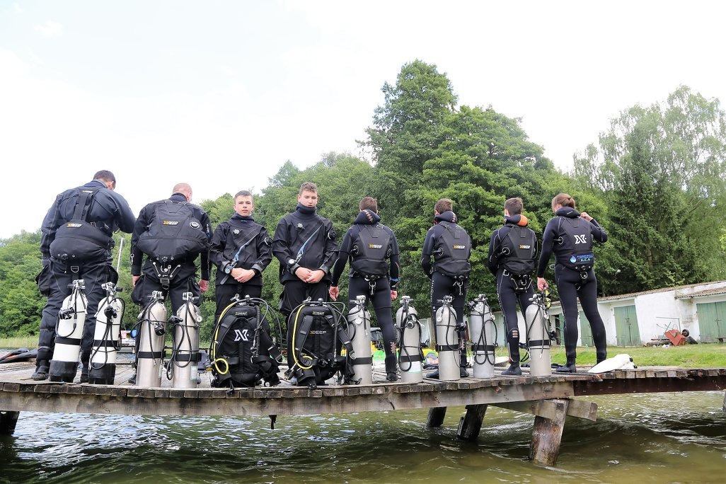 Grupa młodych ludzi w czarnych kombinezonach do nurkowania, stojących na drewnianym pomoście nad wodą, z butlami i sprzętem do nurkowania, przygotowujących się do zejścia pod wodę.