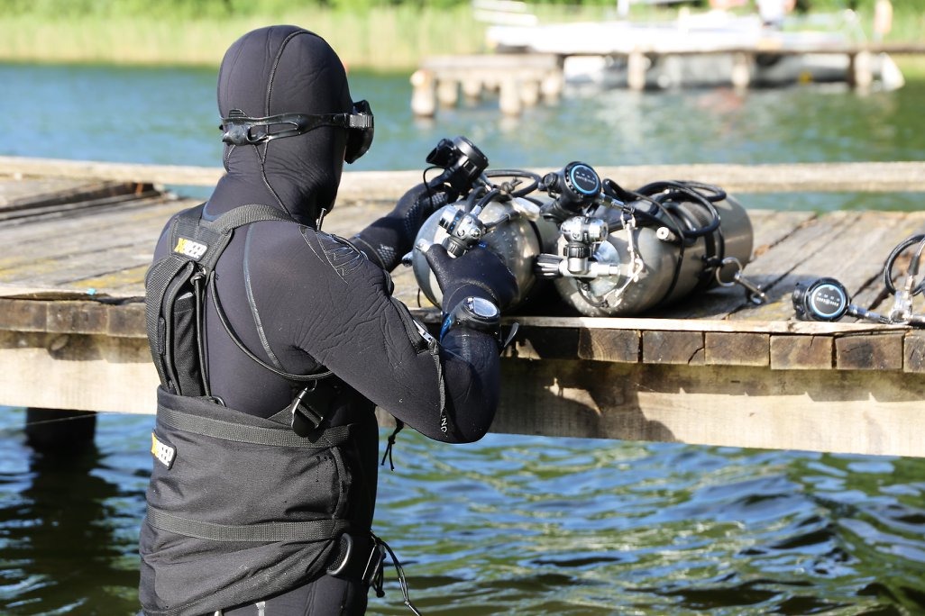 Nurek w czarnym kombinezonie i masce, przygotowujący sprzęt do nurkowania (dwa zbiorniki tlenowe) na drewnianym pomoście nad jeziorem, widok z tyłu.