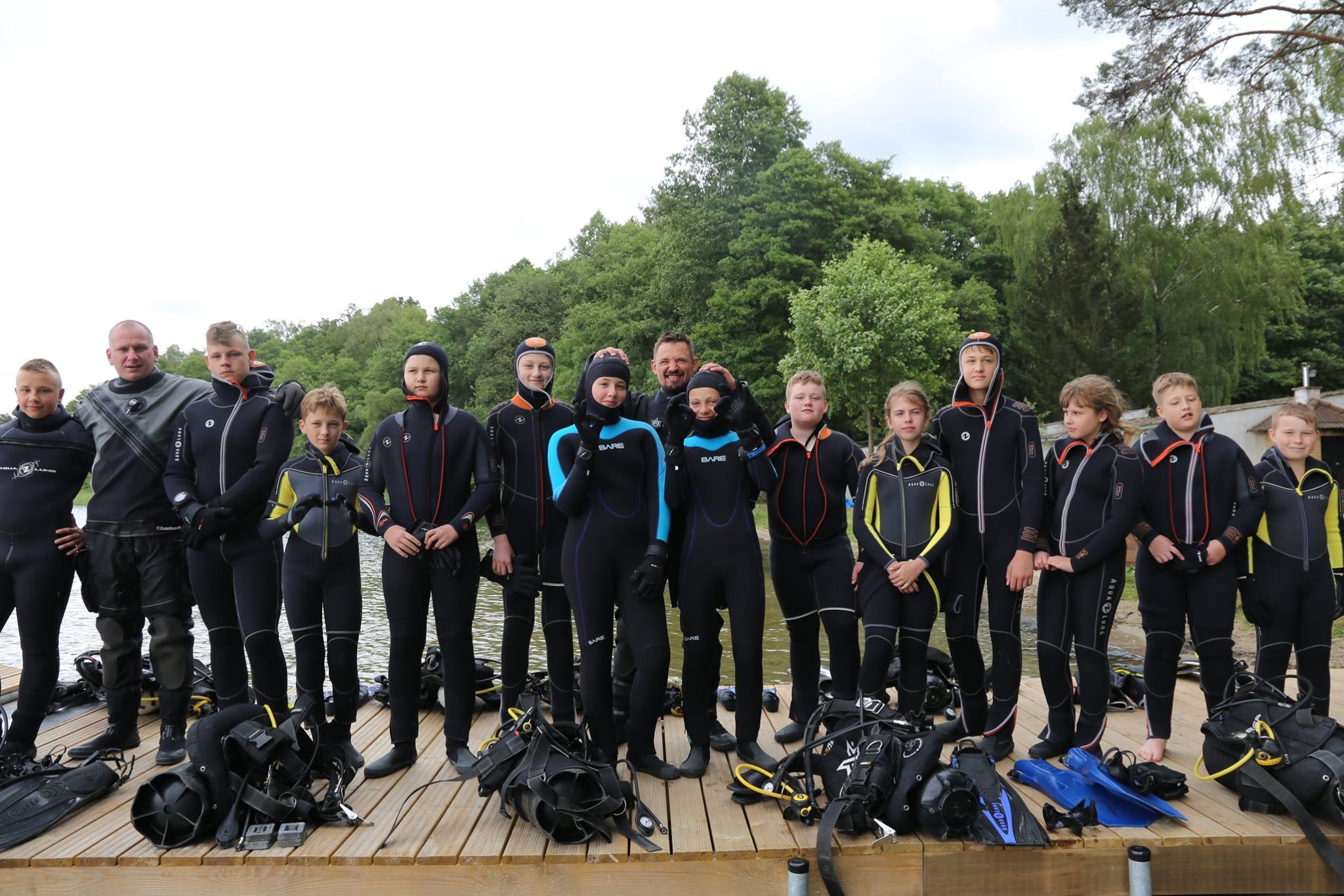 Grupa dzieci i dorosłych w czarnych piankach do nurkowania, stojąca na drewnianym pomoście nad jeziorem, z zielonym lasem w tle i sprzętem do nurkowania u stóp, w tym płetwy i butle.