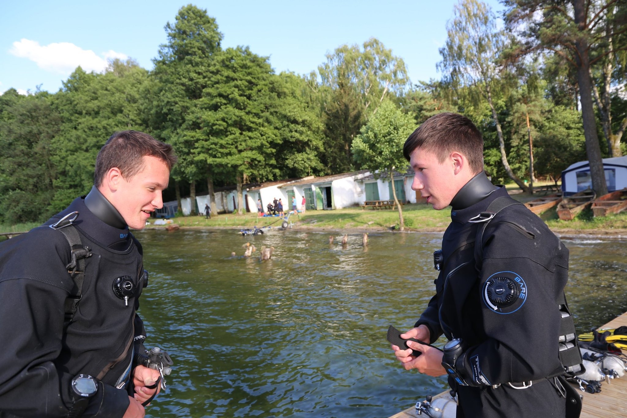 Dwóch nurków w pełnym kombinezonie rozmawia na pomoście nad jeziorem, w tle widać grupę osób w wodzie i budynki.