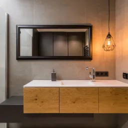 Nowoczesna łazienka z drewnianą szafką pod umywalkę, białym blatem, czarnym lustrem w ramie i geometryczną lampą wiszącą, widoczny fragment kabiny prysznicowej.