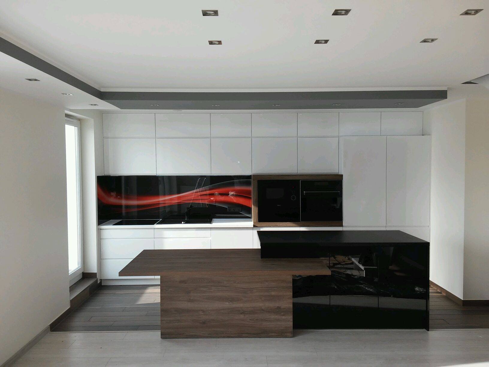 Nowoczesna kuchnia z białymi szafkami, czarnym blatem i wyspą z elementami drewnopodobnymi oraz czarnym panelem ściennym z abstrakcyjnym czerwonym wzorem.