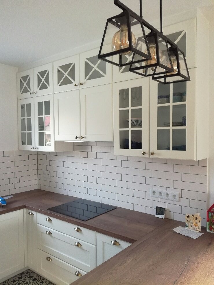 Biała kuchnia z drewnianym blatem, górne szafki z przeszklonymi frontami i dekoracyjnymi krzyżami, czarna lampa wisząca w stylu industrialnym