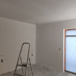 Adrian-bud - Wnętrze w trakcie remontu: świeżo pomalowane ściany na szaro, aluminiowa drabina, zabezpieczone folią drzwi, widoczne gniazdka elektryczne bez osprzętu.