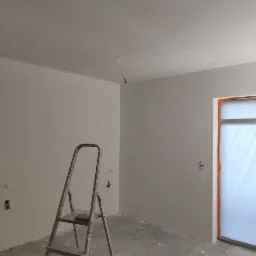 Wnętrze w trakcie remontu: świeżo pomalowane ściany na szaro, aluminiowa drabina, zabezpieczone folią drzwi, widoczne gniazdka elektryczne bez osprzętu.