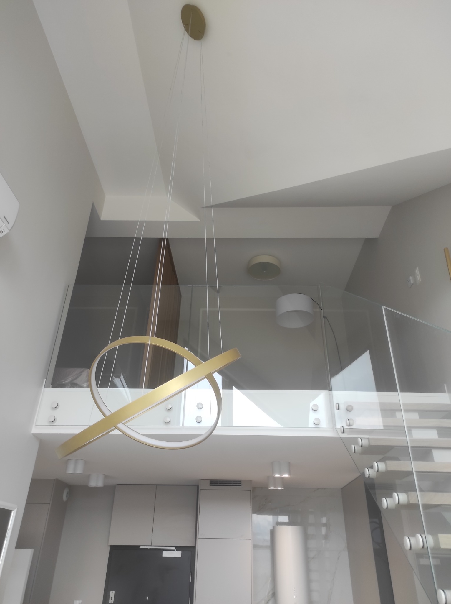 Nowoczesny, dwupoziomowy salon z szklaną balustradą na antresoli i minimalistycznym, złotym, okrągłym żyrandolem wiszącym na cienkich linkach.