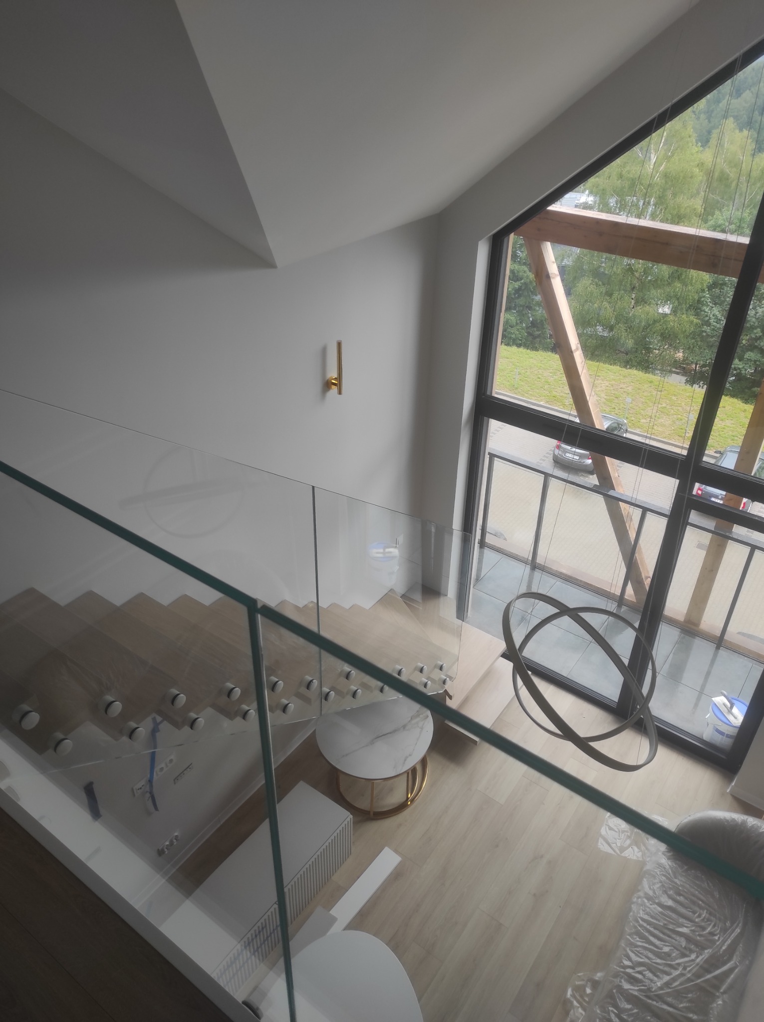 Nowoczesne wnętrze z przeszkloną balustradą schodów, drewnianymi stopniami zabezpieczonymi folią, widokiem na zewnątrz przez duże okno z drewnianą konstrukcją oraz okrągłą lampą wiszącą.