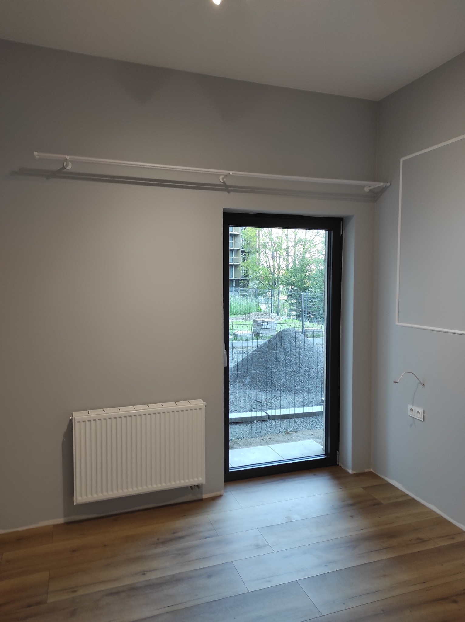 Szare wnętrze pokoju z drewnianą podłogą, białym grzejnikiem i oknem balkonowym z widokiem na plac budowy.