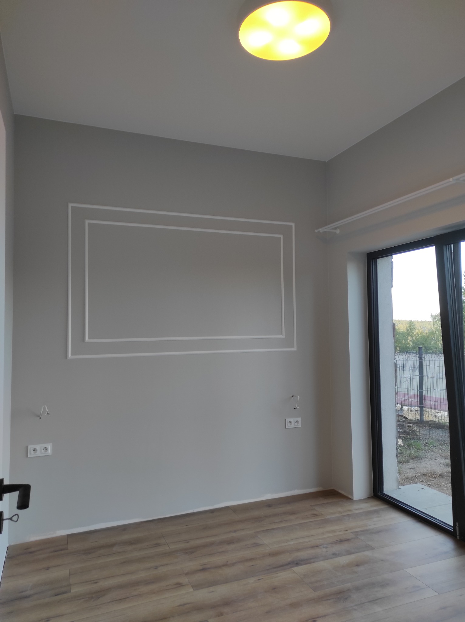 Szare wnętrze pokoju z panelami podłogowymi, białym gzymsem na ścianie, gniazdkami elektrycznymi i drzwiami balkonowymi z widokiem na zewnątrz.